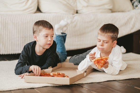two boys lie on the floor eating pepperoni pizza from a box.