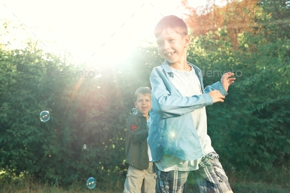 boy against the background of the sun runs after soap bubbles. Happy childhood of two brothers