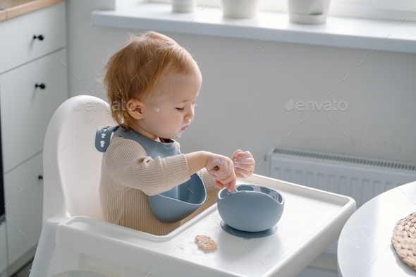 A grubby toddler sitting in a highchair, self feeding, messy eating, learning to eat.