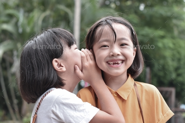 Two little sister girls whisper in ear at park outdoor.