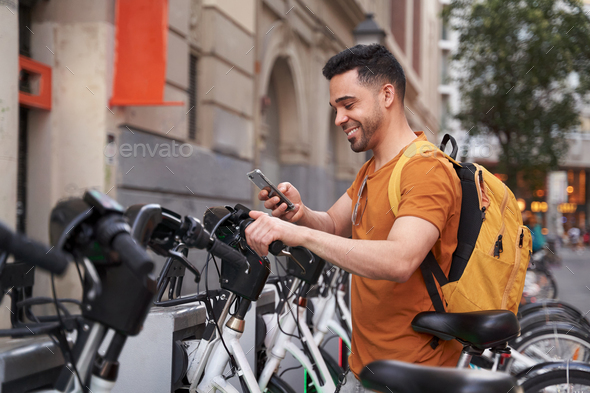 Positive young Latino man stands next to parking lot with rental bike and uses smartphone