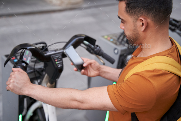 Young Latino man stands by the parking lot with a rental bike and uses a smartphone