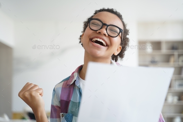 Happy mixed race girl student makes yes gesture, holding document, celebrating success, achievement