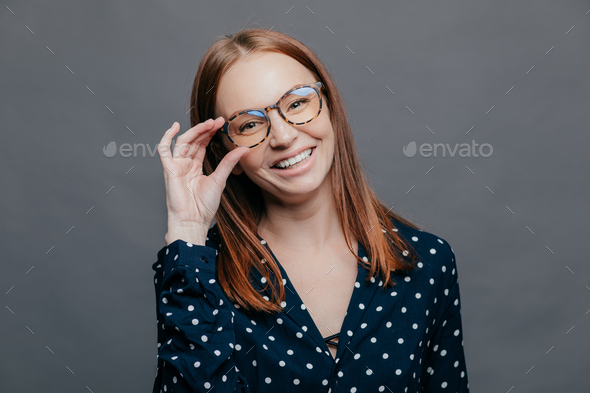 Smiling woman with pleased facial expression, keeps hand on rim of spectacles