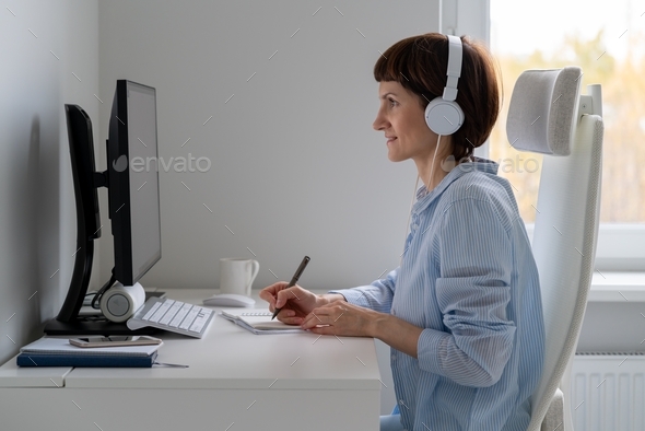Happy female in wireless headphones studying online course, using pc and writing in notepad,