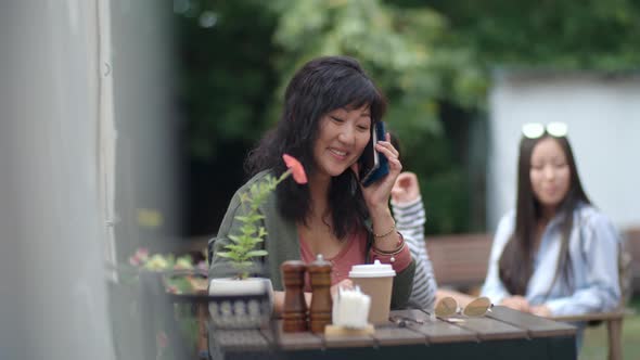 Cheerful Asian Woman Sitting in Outdoor Restaurant and Chatting on Cell Phone alt