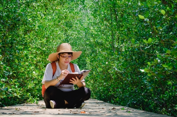 Female traveler taking notes while sitting on walkway in mangrove forest at natural parkland