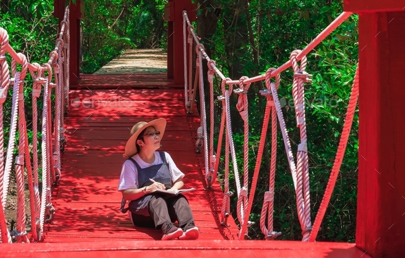 Female tourist taking note while sitting on suspension bridge in mangrove forest at natural parkland