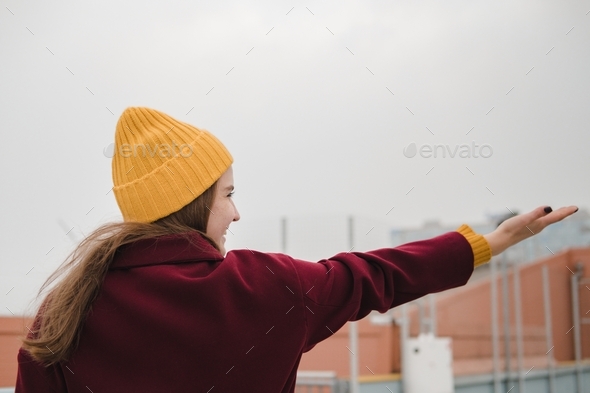 Young beautiful woman in burgundy coat and yellow hat walks along the roof of the parking lot.