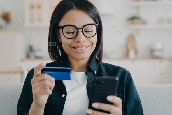 Happy female holding credit card, smartphone, uses online banking services, mobile bank app at home