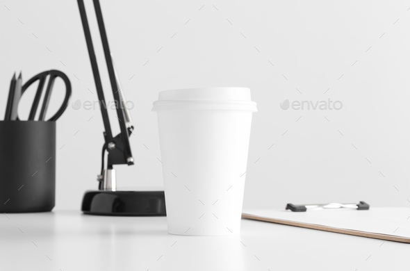 Coffee paper cup mockup with a lamp, clipboard and workspace accessories on a white table.