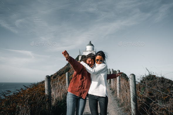 two caucasian young women hugging happily on the narrow dirt road by the fence near the lighthouse