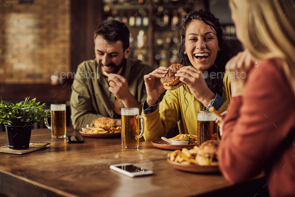 Happy woman having fun while eating hamburger with her friends in a pub.
