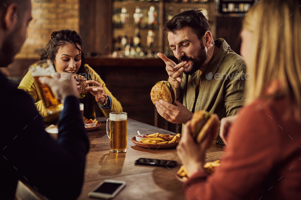 Young happy man eating hamburger with his friends in a pub.