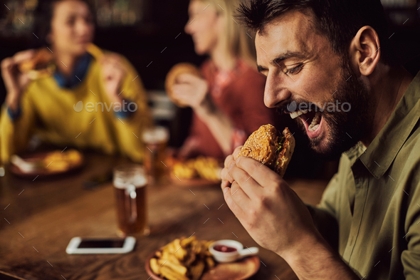 Young man enjoying in hamburger while eating with friends in a pub.