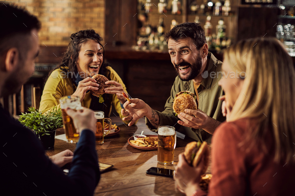 Group of cheerful friends talking while eating hamburgers in a pub.