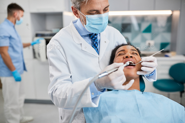 Dentist using dental drill while treating Black woman\'s teeth at dental clinic.
