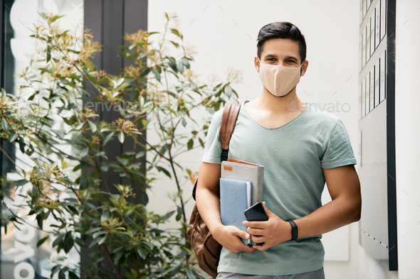 Portrait of happy university student with face mask standing in hallway and looking at camera.