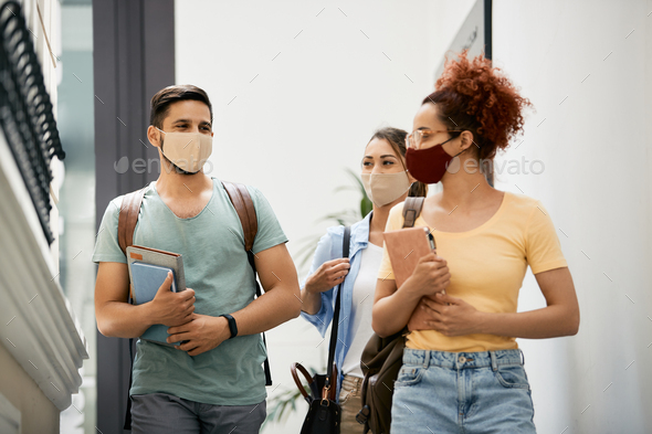 Group of happy university students with face masks talking while walking through hallway.