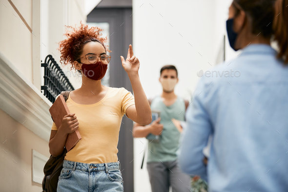 Happy college student with face mask greeting her friend while walking through university hallway.