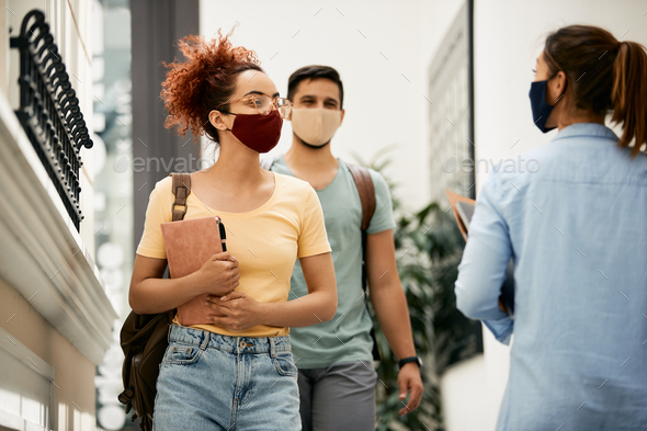 Happy female student with protective face mask greeting her friend at university hallway.