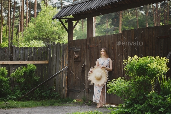 A girl in a long dress stands at the wooden gate of a country house
