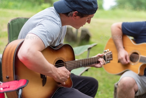 Two guys playing acoustic guitars at a campsite