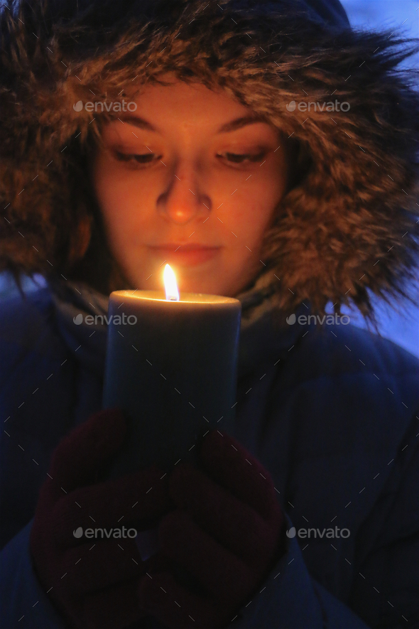 Vertical portrait of illuminated young woman wearing fur trimmed parka holding lighted candle.