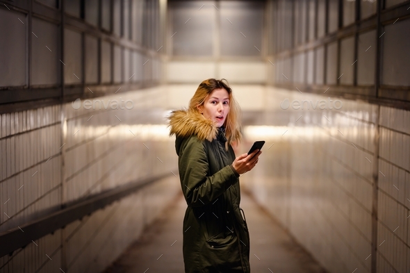 Young blonde woman in green winter parka with fur looks at smartphone in underground crossing