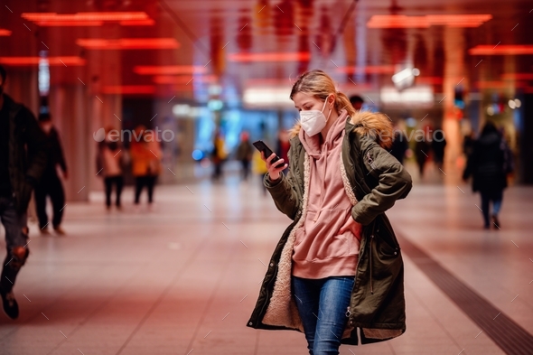 Young blonde woman in protective mask with ponytail in green fur parka surfing through smartphone