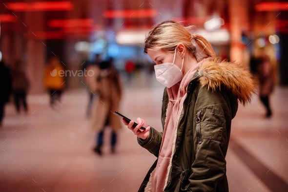 Young blonde woman in protective mask with ponytail in green fur parka surfing through smartphone