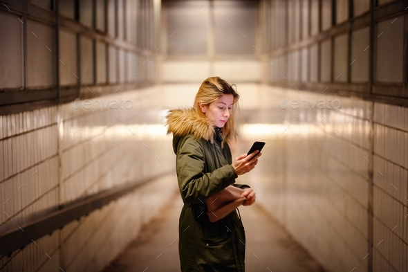 Young blonde woman in green winter parka with fur looks at smartphone in underground crossing