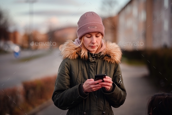 Young blonde girl in hat and green fur parka using smartphone on street