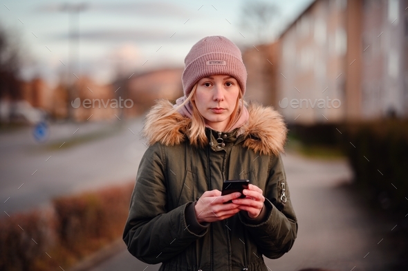 Young blonde girl in hat and green fur parka using smartphone on street