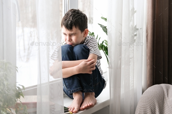 A sad boy sits on the windowsill hugging his knees. Bad mood, depression