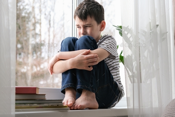 A sad boy sits on the windowsill hugging his knees. Bad mood, depression.