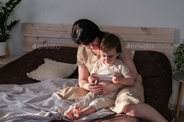 Mom and baby watch cartoons on a smartphone while sitting on the bed