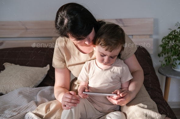 Mom and baby watch cartoons on a smartphone while sitting on the bed