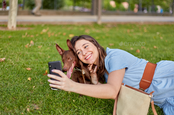 Pretty young woman taking a selfie with her pharaoh dog lying on the grass in a park.