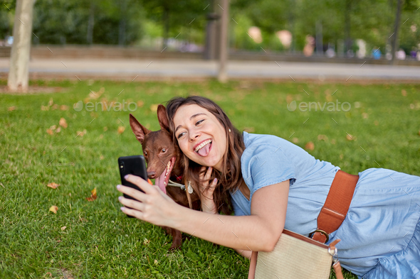 Woman sticking out her tongue taking a selfie with her dog Pharaoh lying on the grass in a park.
