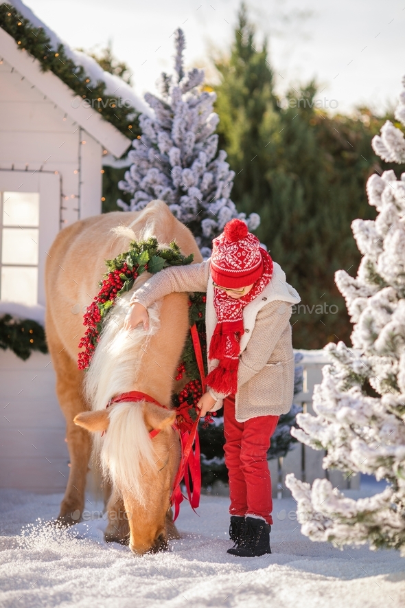 Little boy with glasses holds his adorable pony with festive wreath near the small wooden house