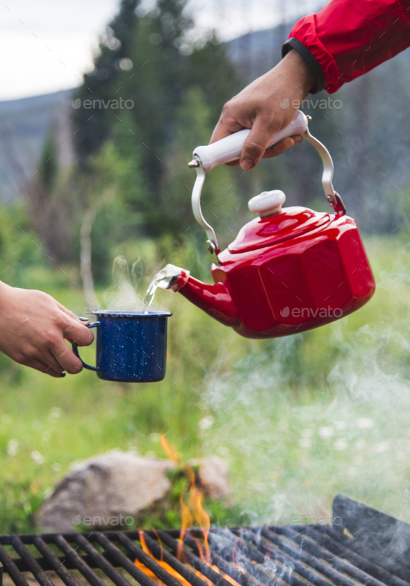Man Pouring hot water from a red kettle into a blue coffee mug at campsite in the morning near fire