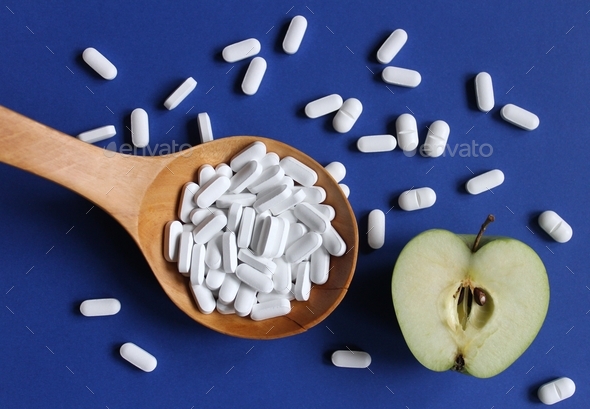 Healthy pills in a wooden spoon and half of apple fruit on blue background