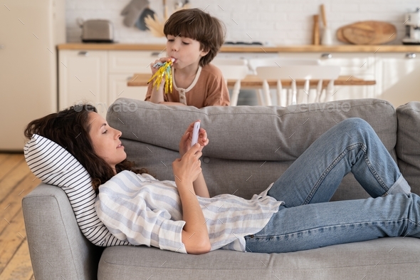 Calm mom lying on sofa with smartphone distracted from noisy loud son bad behavior drawing attention