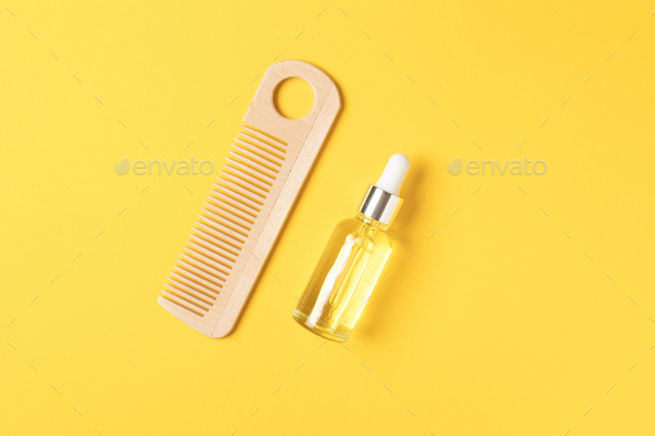 Natural hair care oil and wooden comb on yellow background, top view.