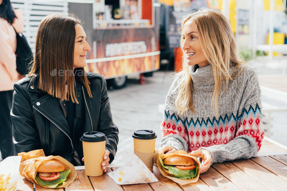 Two happy hungry female friends are chatting and eating burgers at the street market.