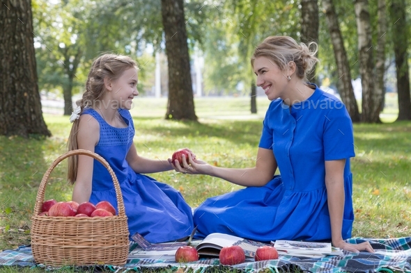 Caucasian mom and teenage daughter are resting in the park on a blanket with a basket of red apples.