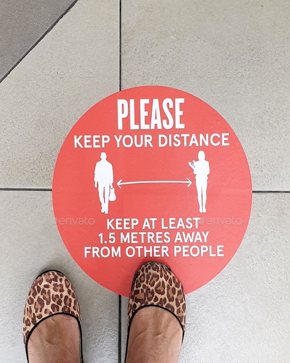 Lower section of woman standing above red social distancing safety sign on floor