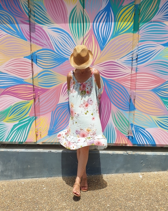 woman wearing summer dress with hat standing against colourful mural art on wall