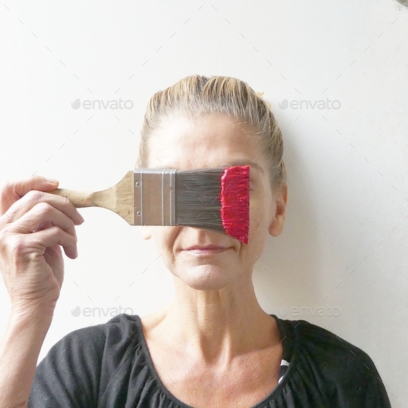 Woman holding paintbrush with red paint obscuring face indoors against white wall